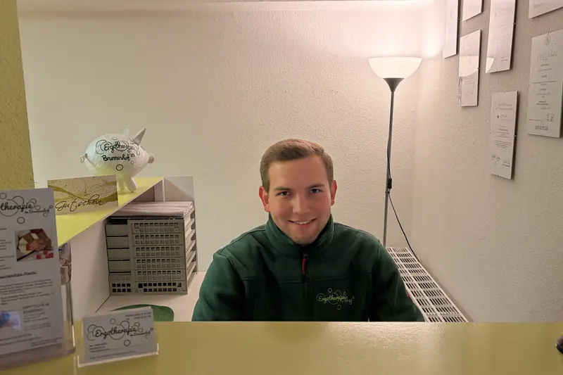 Ein Mitarbeiter in grüner Praxisjacke am Empfangstresen der Praxis, daneben Flyer der Ergotherapie im Brunnenhof