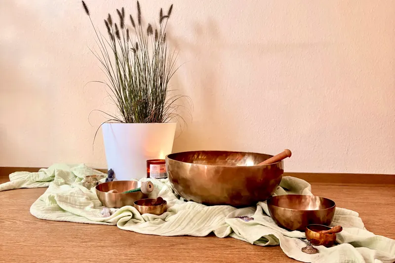 Große und kleine Klangschalen auf einem hellgrünen Tuch vor einer Ziergraspflanze, daneben eine brennende Kerze