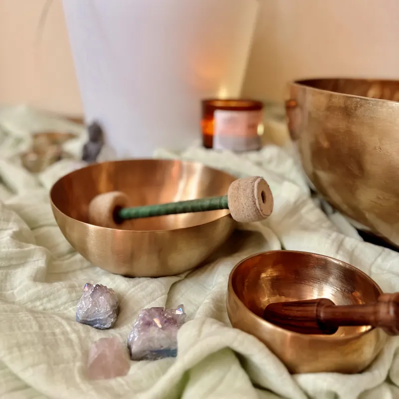 Klangschalen aus Bronze mit Filzschlägel auf einem hellgrünen Tuch, davor Amethyst-Steine, im Hintergrund eine Kerze